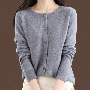 Puede incluir: Un cárdigan gris con cuello redondo y cierre de botones. El suéter de manga larga está hecho de un material de punto suave. Los botones son de color gris a juego.