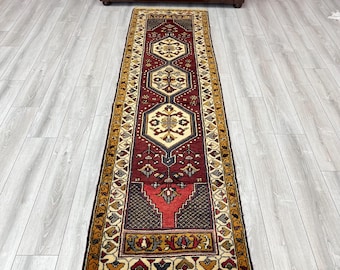 May include: A long, rectangular rug with a detailed geometric pattern. The rug features a deep red central field with cream, gold, and navy blue accents. The border is a mix of gold and cream colors, creating a traditional aesthetic.