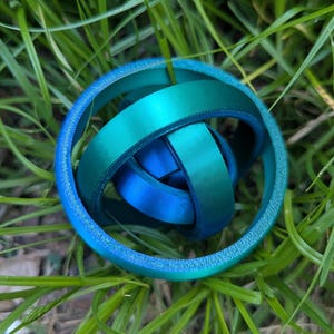 May include: A teal and blue interlocking ring sculpture rests in green grass. The sculpture has a gradient effect, transitioning from teal to blue. The rings are smooth and appear to be made of a solid material.