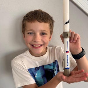 May include: A white model rocket with a pointed nose cone, featuring a black feather graphic, a small American flag, and the text "NEW GLENN" and "BLUE ORIGIN" printed in blue on the cylindrical body, with metallic-colored fins at the base.