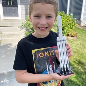 Può includere: Un modello di razzo bianco e nero con una base nera è tenuto in mano da una persona che indossa una maglietta nera con la scritta "IGNITE" e una grafica di navetta spaziale. Il razzo ha più cilindri bianchi e dettagli neri.