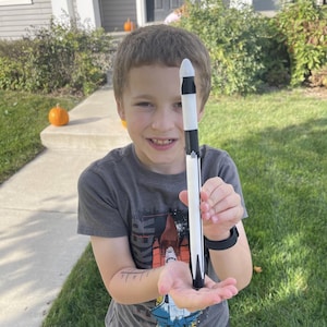 May include: A black, white, and silver toy rocket held by a person. The rocket has a pointed nose cone and a detailed design. The person is wearing a gray t-shirt with a graphic of a space shuttle. The background includes a house, pumpkins, and green grass.