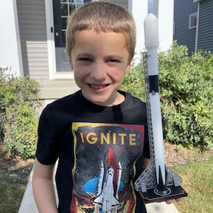 May include: A black t-shirt with the words "IGNITE YOUR IMAGINATION" and a graphic of a space shuttle launching. A white, black, and grey toy rocket is held in front of the shirt.