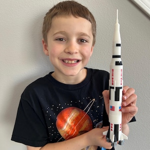 May include: A white Saturn V rocket model featuring black detailing, the text "UNITED STATES" in red, and small American flag decals. The rocket has a pointed top, a cylindrical body, and a multi-engine base with four stabilizing fins at the bottom.