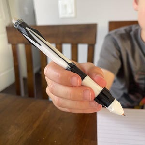 May include: A white and black pen with a black tip and eraser. The pen is held by a person over a sheet of lined paper on a wooden table. The pen has a unique design.