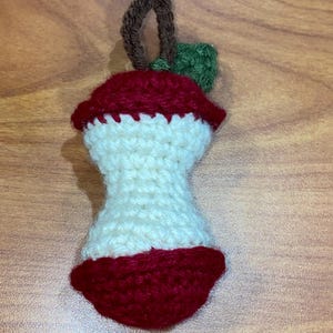 May include: A crocheted apple core ornament in red, white, and green. The apple core has a red top and bottom, with a white centre. A brown loop and green leaf are attached to the top. Ideal for autumn decorations.