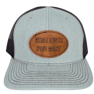 Puede incluir: Gorra trucker gris claro con parte trasera de malla y visera negras. La gorra presenta un parche de cuero marrón con las palabras "WHAT EVER YOU WANT" en letras negras. La visera tiene costuras negras.