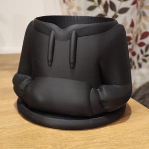 May include: A black planter shaped like a hoodie, complete with a hood, drawstrings, and a front pocket. The planter sits on a round base. The design is modern and minimalist, suitable for small plants or succulents.
