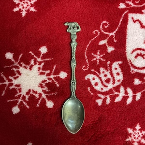 May include: A silver-colored spoon with a decorative handle featuring a bear figure. The spoon has an oval bowl and ornate detailing on the handle. The spoon is set against a red background with white snowflake and floral patterns.