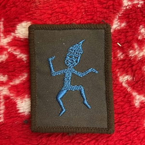 Könnte beinhalten: Rechteckiger Aufnäher mit schwarzem Hintergrund und einer blau gestickten Figur. Die Figur hat einen spitzen Hut und ausgestreckte Arme und Beine. Der Aufnäher hat einen braunen Rand und ist vor einem roten, strukturierten Hintergrund platziert.