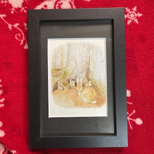 Könnte beinhalten: Gerahmte Illustration mit einer Waldszene mit Kaninchen in der Nähe eines Baumes. Das Kunstwerk befindet sich in einem schwarzen Rahmen, vor einem roten Hintergrund mit weißem Muster. Die Illustration hat weiche, gedämpfte Farben.
