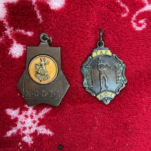 May include: Two vintage medals with ornate designs. One is a bronze-toned pendant with a dancer in a yellow circle, and the letters N.C.D.T.A. The other is a silver-toned medal with a dancer and the letters T.A.P. A.D.A.