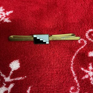 Op de afbeelding: Een goudkleurige stropdasklem met een rechthoekig ontwerp met een gepixeld zwart, wit en grijs patroon. De clip staat tegen een rode, getextureerde achtergrond met witte sneeuwvlokachtige patronen.