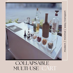 Puede incluir: Un carrito multiusos plegable blanco con suministros de bar. El carrito tiene un estante de acrílico transparente con vasos de fruta seca y botellas. Se muestra el texto "COLLAPSIBLE MULTI-USE CART".