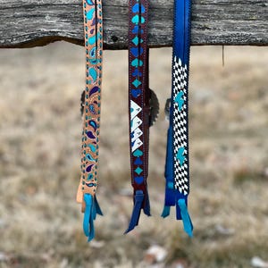 May include: Three leather horse tack straps with decorative designs. The straps are tan, brown, and blue, each featuring unique patterns in turquoise, white, and blue. Each strap has a leather fringe at the bottom. The straps are hanging on a wooden fence.