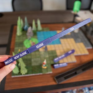 May include: A purple game range ruler with white text indicating "MELEE VERY CLOSE", "CLOSE", and "FAR". The ruler is held over a miniature tabletop game with trees, water, and other game pieces.