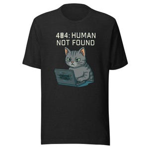 Puede incluir: Camiseta gris oscuro con un gato de dibujos animados usando una computadora portátil. El gato es gris con rayas negras y tiene una expresión de mal humor. El texto "404: HUMAN NOT FOUND" está encima del gato en blanco. La camiseta es de material suave.