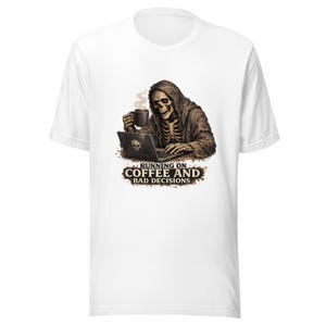 May include: White t-shirt with a graphic of a skeleton drinking coffee and using a laptop. The text "RUNNING ON COFFEE AND BAD DECISIONS" is below the graphic. The design is in shades of brown and black.