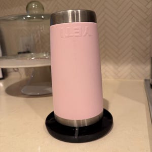 May include: A pink insulated tumbler with a stainless steel rim and lid, resting on a black coaster. The word "YETI" is printed on the side of the tumbler. The tumbler is cylindrical in shape.