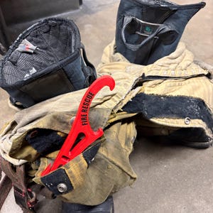 Puede incluir: Equipo de bombero, que incluye un abrigo beige y negro, botas y una herramienta roja "FIRE RESCUE". El abrigo muestra signos de desgaste. Las botas son negras. El equipo está sobre un suelo de hormigón.