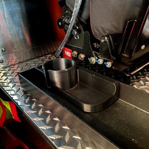 May include: A black plastic cup holder and tray combo, designed to fit within a vehicle's interior. The cup holder is cylindrical, and the tray is rectangular with rounded corners. The items are set against a backdrop of a vehicle seat and diamond-plate metal.