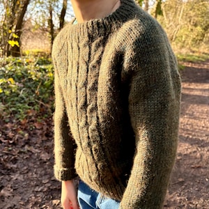 Può includere: Un maglione verde a trecce. Il maglione ha un girocollo e maniche lunghe. Il motivo a trecce è visibile sul davanti. Il maglione è realizzato con un filato spesso e strutturato. La persona indossa jeans blu.