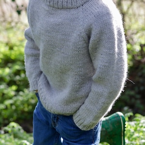 May include: A pale grey knitted jumper with a crew neck. The jumper is paired with blue jeans. A green wellington boot is visible in the lower right corner. The background is a blurred outdoor setting.