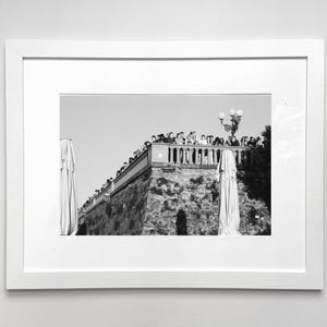 Könnte beinhalten: Schwarz-weiß gerahmtes Foto einer Steinmauer mit einer Gruppe von Menschen oben. Die Mauer hat ein dekoratives Geländer und eine Straßenlaterne. Zwei geschlossene Sonnenschirme sind sichtbar.
