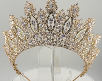 May include: A gold-coloured tiara with a symmetrical design. The crown features clear, sparkling crystals and a delicate, ornate structure. The tiara is displayed on a clear stand, showcasing its intricate details and elegant design.