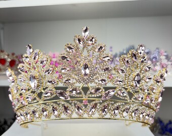 May include: A gold-toned crown with clear and pink crystals. The tiara features a symmetrical design with large, teardrop-shaped crystals and smaller, round stones. The crown is likely for a special occasion, such as a wedding or pageant.
