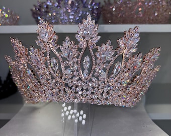 May include: A rose gold tiara with clear, sparkling crystals. The crown features a leaf-like design with large, central crystals and smaller stones throughout. Other tiaras are visible in the background, displayed on a shelf.