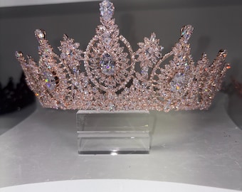 May include: A rose gold tiara with clear, sparkling crystals. The crown features a symmetrical design with a central, large crystal and smaller crystals throughout. The tiara is displayed on a clear stand, showcasing its intricate details.