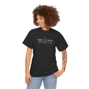 May include: Black t-shirt with the text "Home is where 127.0.0.1 is" in white. The shirt is a crew neck style. The person wearing the shirt is smiling and wearing blue jeans.