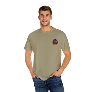 Pode incluir: Uma camiseta verde-oliva clara com um pequeno emblema circular na parte superior esquerda do peito. O emblema apresenta um dragão vermelho e texto em um design circular. A pessoa está usando jeans azul.