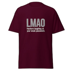 Può includere: T-shirt bordeaux con la scritta "LMAO" in un font pixelato, sopra la frase "Gli hacker ridono della tua password debole". La maglietta è tinta unita e ha maniche corte.