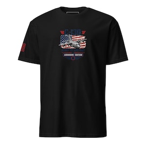 May include: A black short-sleeve t-shirt featuring a graphic of a C-130 aircraft over a distressed American flag. The text reads C-130, HURRY UP AND WAIT, and AIRBORNE EDITION. A small American flag graphic is printed on the right sleeve.