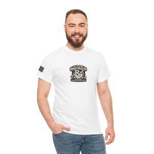 May include: White t-shirt with a graphic that reads "THE ORIGINAL DRUG DEALER" above a stylized "Rx" symbol and the text "LICENSED AND EVERYTHING". The shirt has a crew neck and short sleeves. The person is wearing blue jeans.