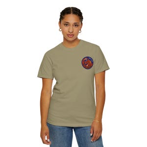 May include: A tan crew neck t-shirt with a circular patch on the left chest. The patch features a red, white, and blue design with text. The model is wearing blue jeans.