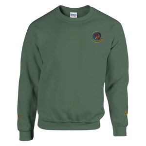 May include: A green crewneck sweatshirt with a small emblem on the upper chest. The emblem features a red dragon and text. The sleeve has embroidered text. The sweatshirt is made of a soft material.