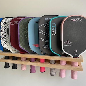 May include: A wooden rack displaying seven pickleball paddles with various designs and colors. The paddles are held in place by a wooden rack with holes for the handles. The paddles have black, blue, red, and pink accents.