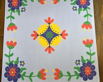 May include: A square light blue mat featuring a central diamond-shaped floral motif in yellow, red, and dark blue. The corners are decorated with large dark blue flowers, while the edges are connected by green vines with red and blue blossoms.