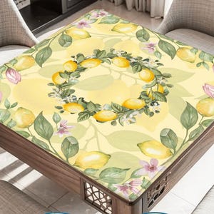 May include: A square table with a yellow lemon-themed tablecloth. The tablecloth features a lemon wreath design in the center, surrounded by lemons, leaves, and flowers. The table is made of wood and has decorative cutouts.