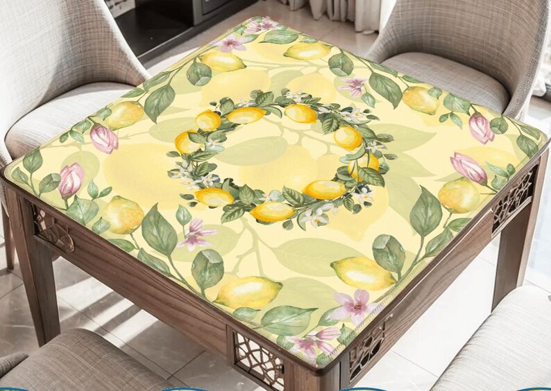 May include: A square table with a yellow lemon-themed tablecloth. The tablecloth features a lemon wreath design in the center, surrounded by lemons, leaves, and flowers. The table is made of wood and has decorative cutouts.