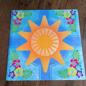 Pu&ograve; includere: Tappeto quadrato blu con un grande disegno di sole arancione al centro. Il sole ha un centro giallo e raggi arancioni. Fiori colorati e foglie verdi decorano gli angoli. Il tappeto ha un tema tropicale.