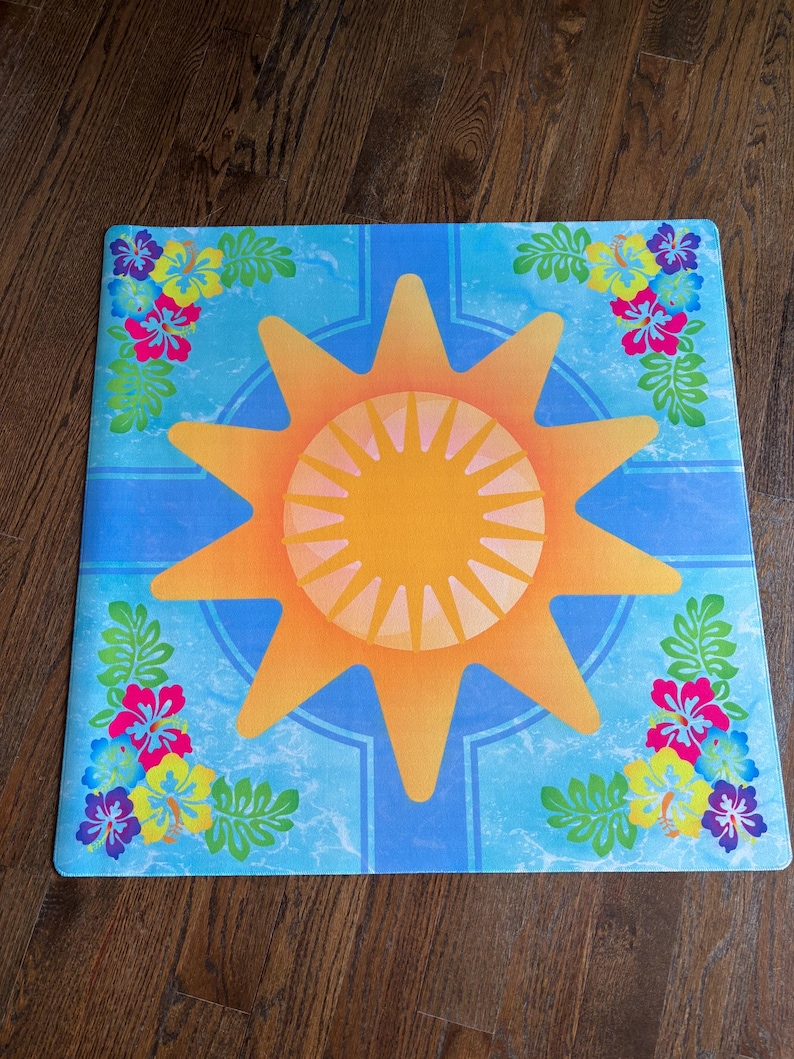 Pu&ograve; includere: Tappeto quadrato blu con un grande disegno di sole arancione al centro. Il sole ha un centro giallo e raggi arancioni. Fiori colorati e foglie verdi decorano gli angoli. Il tappeto ha un tema tropicale.