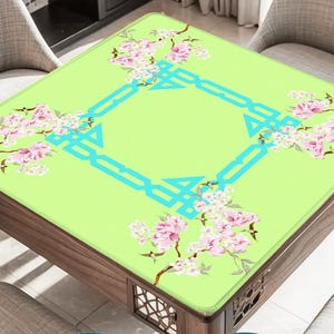 K&ouml;nnte beinhalten: Ein quadratischer Tisch mit einer hellgr&uuml;nen Oberfl&auml;che, verziert mit rosa Bl&uuml;tenzweigen und einem t&uuml;rkisfarbenen geometrischen Design. Der Tisch hat einen dunklen Holzrahmen und ist von St&uuml;hlen umgeben.