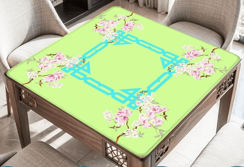 K&ouml;nnte beinhalten: Ein quadratischer Tisch mit einer hellgr&uuml;nen Oberfl&auml;che, verziert mit rosa Bl&uuml;tenzweigen und einem t&uuml;rkisfarbenen geometrischen Design. Der Tisch hat einen dunklen Holzrahmen und ist von St&uuml;hlen umgeben.