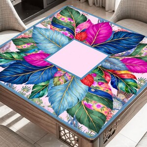 May include: A square table cover featuring a vibrant pattern of large, colorful leaves in shades of blue, green, magenta, and yellow against a light pink background, with a solid light pink square centered in the middle of the design.