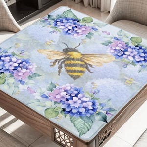 May include: A square table with a floral design featuring blue and purple hydrangeas and a large bee. The table has a wooden frame and is surrounded by chairs. The table's surface is light blue.