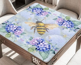 May include: A square table with a floral design featuring blue and purple hydrangeas and a large bee. The table has a wooden frame and is surrounded by chairs. The table's surface is light blue.